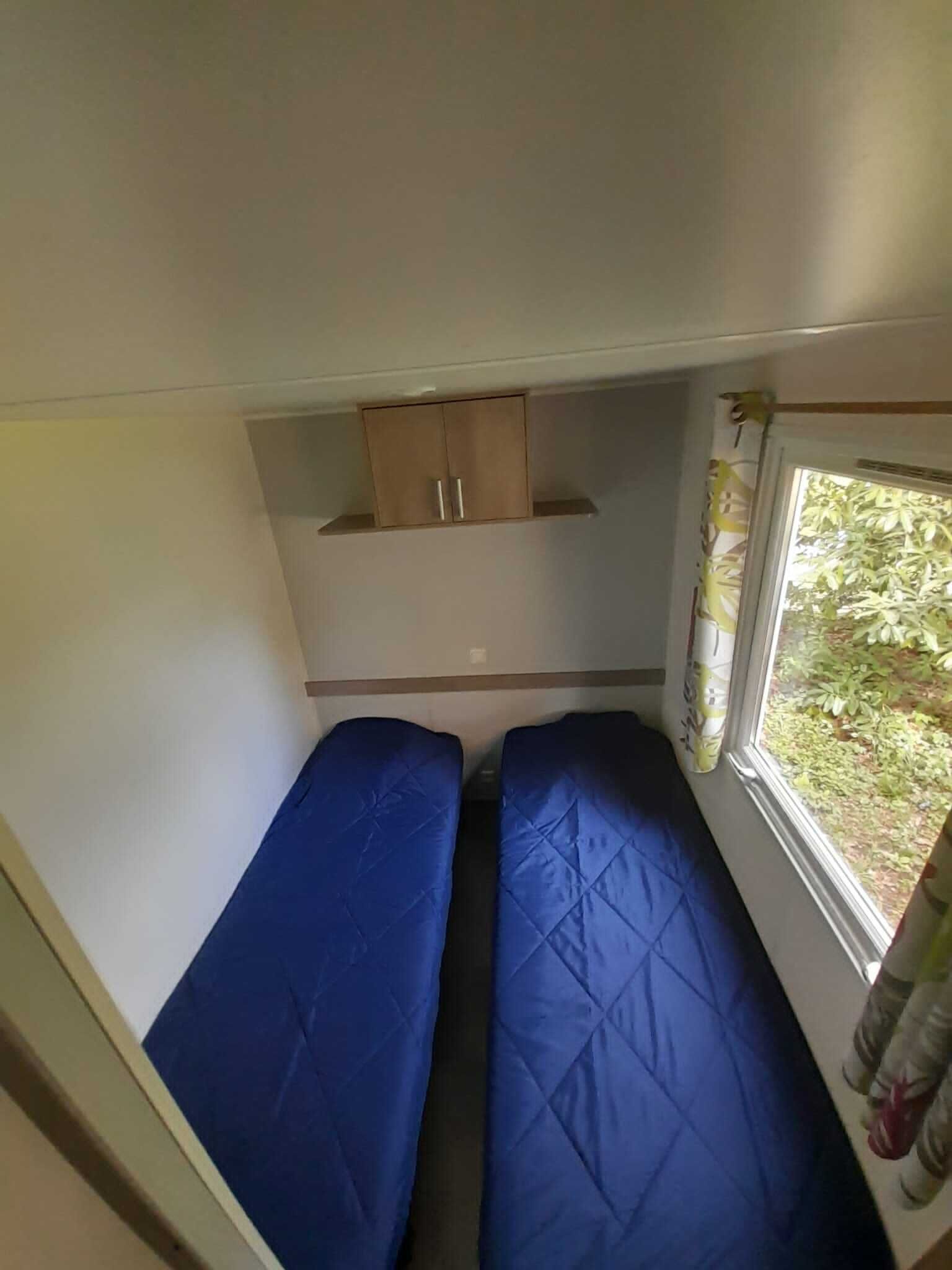 A small, enclosed space with two blue beds, a wooden cabinet, and a window with curtains.