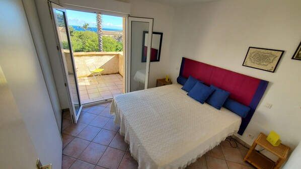 A bedroom with a bed, a nightstand, a window with a view of greenery and a building, and a door leading to an outdoor area.