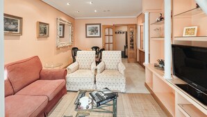 A living room with a sofa, two patterned armchairs, a coffee table with books and a remote control, a television on a stand, and a bookshelf. - Apartamento ThatsHome Silgar 4 (Sanxenxo)