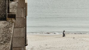 A beach with a person walking a dog, a boat in the water, and a stone building in the foreground. - Apartamento ThatsHome Silgar 4 (Sanxenxo)