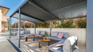 An outdoor patio area with a covered seating arrangement, including sofas and a coffee table, and a view of a garden with autumn foliage. - Brand new 6 bedroom villa, only 1500m from the beach, village Valbandon (Valbandon)