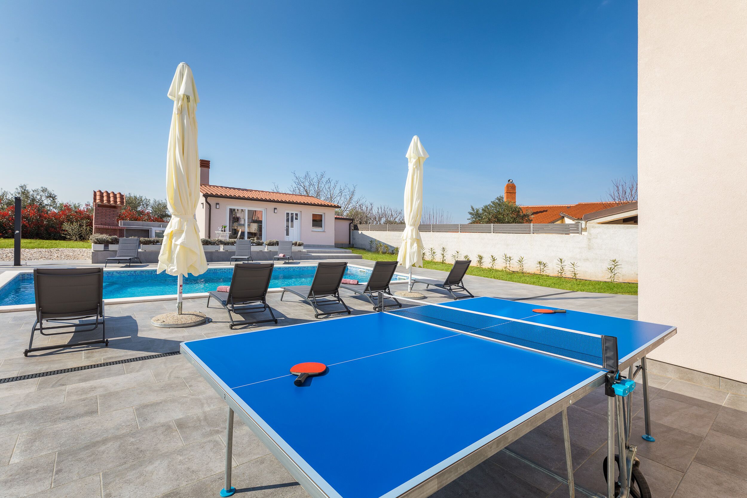 Una mesa de ping pong al aire libre con cuatro sillas y dos sombrillas, una piscina y un edificio al fondo.