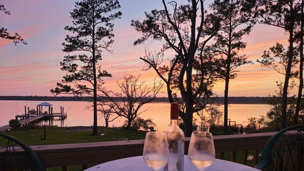 Ein Tisch mit zwei Weingläsern und einer Flasche, vor einer Sonnenuntergangslandschaft mit einer Anlegestelle und Bäumen.
