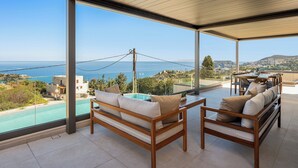 A balcony with a pool, wooden furniture, and a view of the sea and hills. - Kyma Majo Luxury Sea View Villa 6-Bedroom with Gym,Sauna,Heated Private Pool (Malevizi)