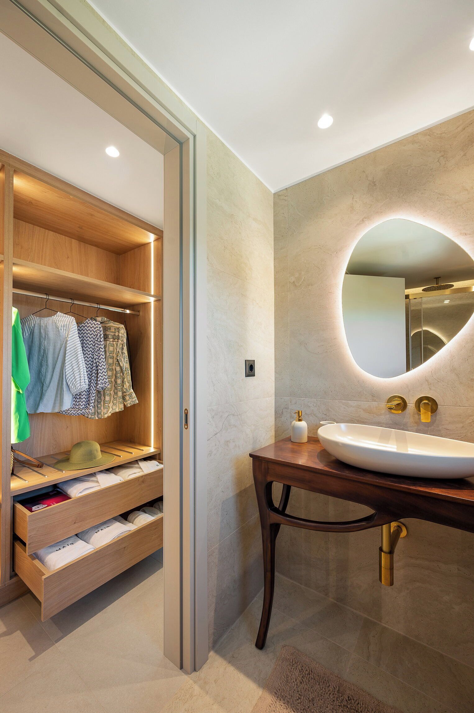 Un baño moderno con tocador de madera, un espejo ovalado grande y un vestidor con ropa colgada y cajones.