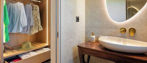 Un baño moderno con tocador de madera, un espejo ovalado grande y un vestidor con ropa colgada y cajones.