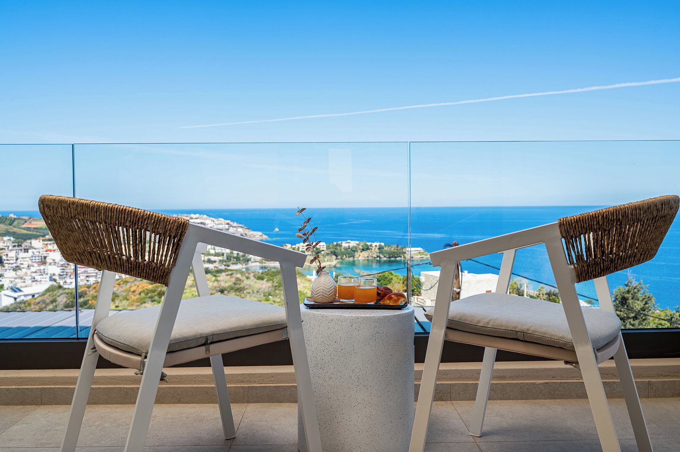 Dos sillas de mimbre con cojines, una pequeña mesa redonda con una bandeja de bebidas y un florero, y una vista panorámica del mar y un pueblo costero.