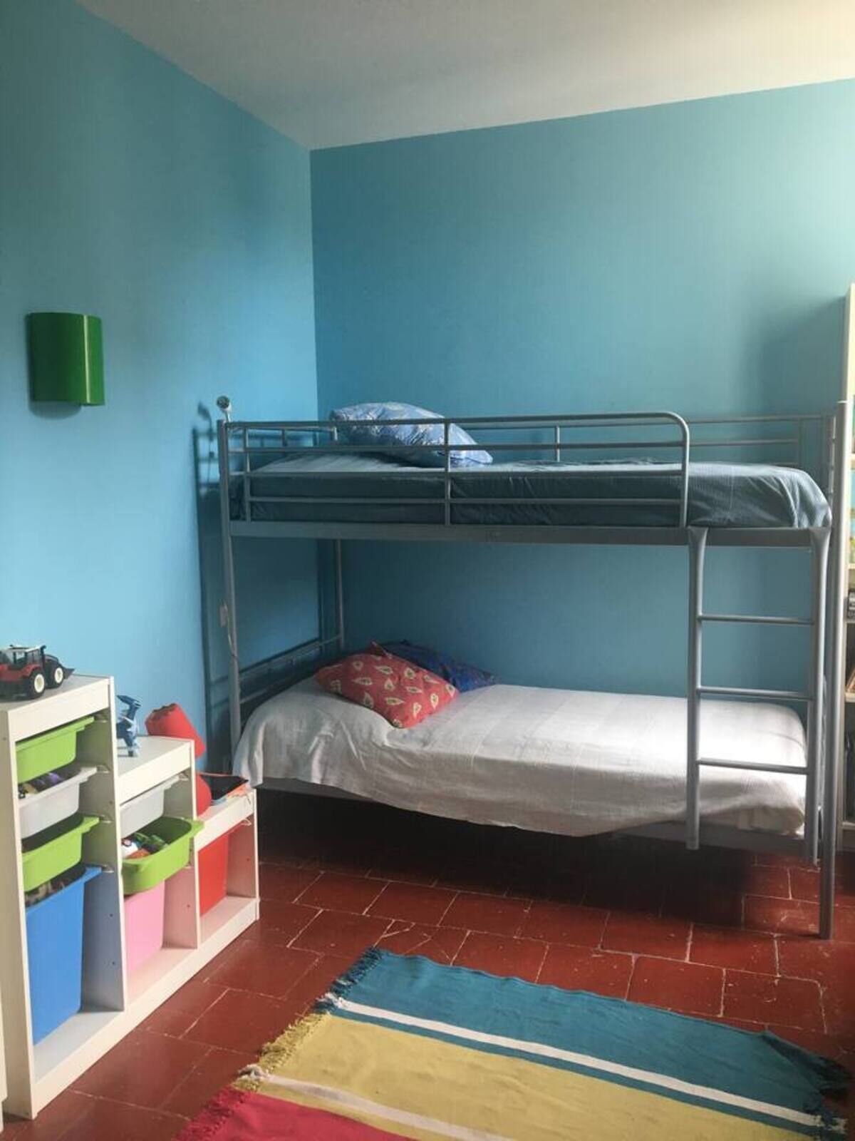 A bunk bed room with a blue wall, a green trash bin, and a colorful rug.