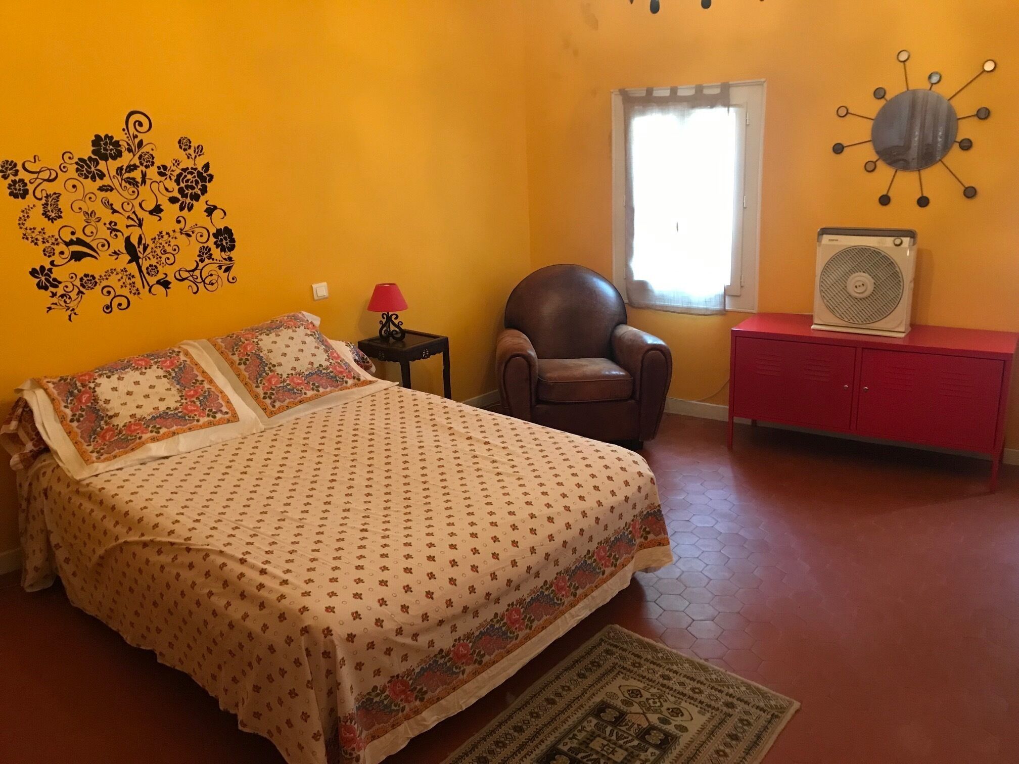 Un dormitorio con una cama, una silla, un armario rojo, un reloj de pared y una ventana con cortinas.