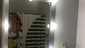 Une salle de bain avec un miroir de coiffeuse entouré de lumières, un rideau de douche et une prise électrique au mur.