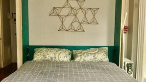 Un lit surmonté d’une œuvre d’art murale aux motifs géométriques, avec une tête de lit verte et des coussins à motifs.