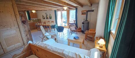 A cozy room with a wooden ceiling, a sofa, a dining table with chairs, a fireplace, and a window with curtains.
