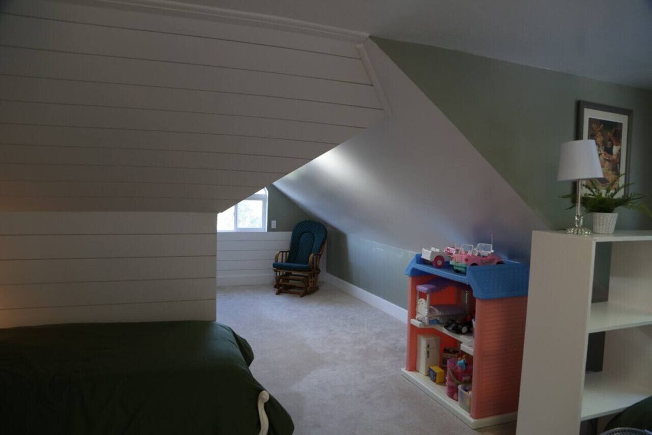 Une chambre avec un plafond en pente, un fauteuil à bascule, une bibliothèque et une fenêtre.