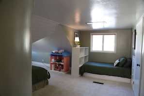 A small, well-lit room with two beds, a window, a shelf with toys, and a door. - Updated Cottage Sleeps 8 Close to Logan (River Heights)