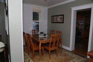 A dining area with a wooden table and chairs, a window with blinds, a framed picture on the wall, and a kitchen visible through an open doorway. - Updated Cottage Sleeps 8 Close to Logan (River Heights)