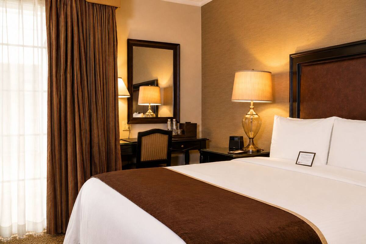 Une chambre d’hôtel avec un grand lit, un bureau avec une chaise, une lampe, un miroir et une vue sur l’extérieur à travers des rideaux transparents.