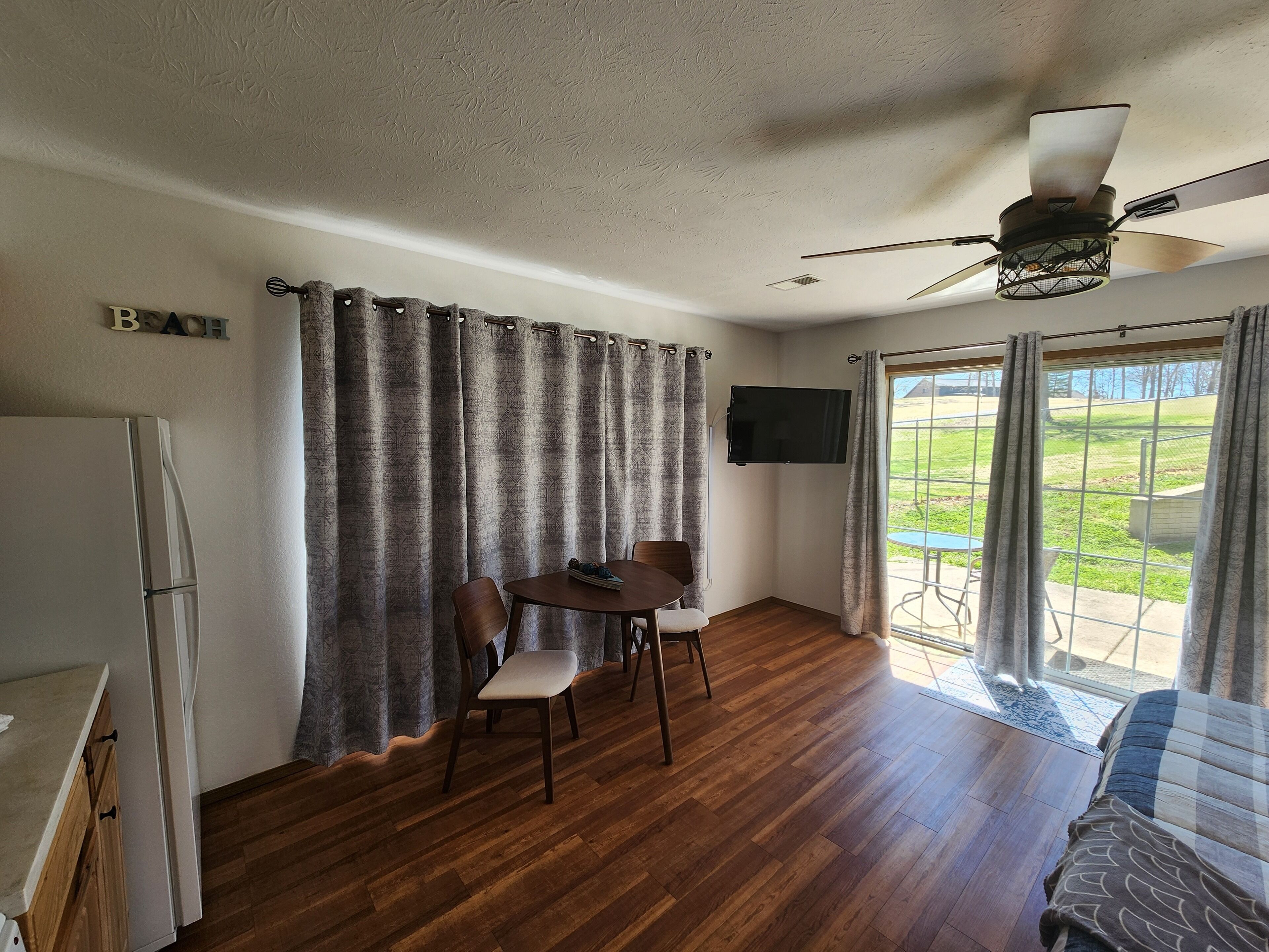 A room with a wooden floor, a dining table with chairs, a ceiling fan, a TV mounted on the wall, and a view of a grassy area through a sliding door.