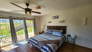 Une chambre à coucher comprenant un lit, un ventilateur de plafond, une table de chevet et une vue sur un espace verdoyant, visible à travers une porte coulissante.
