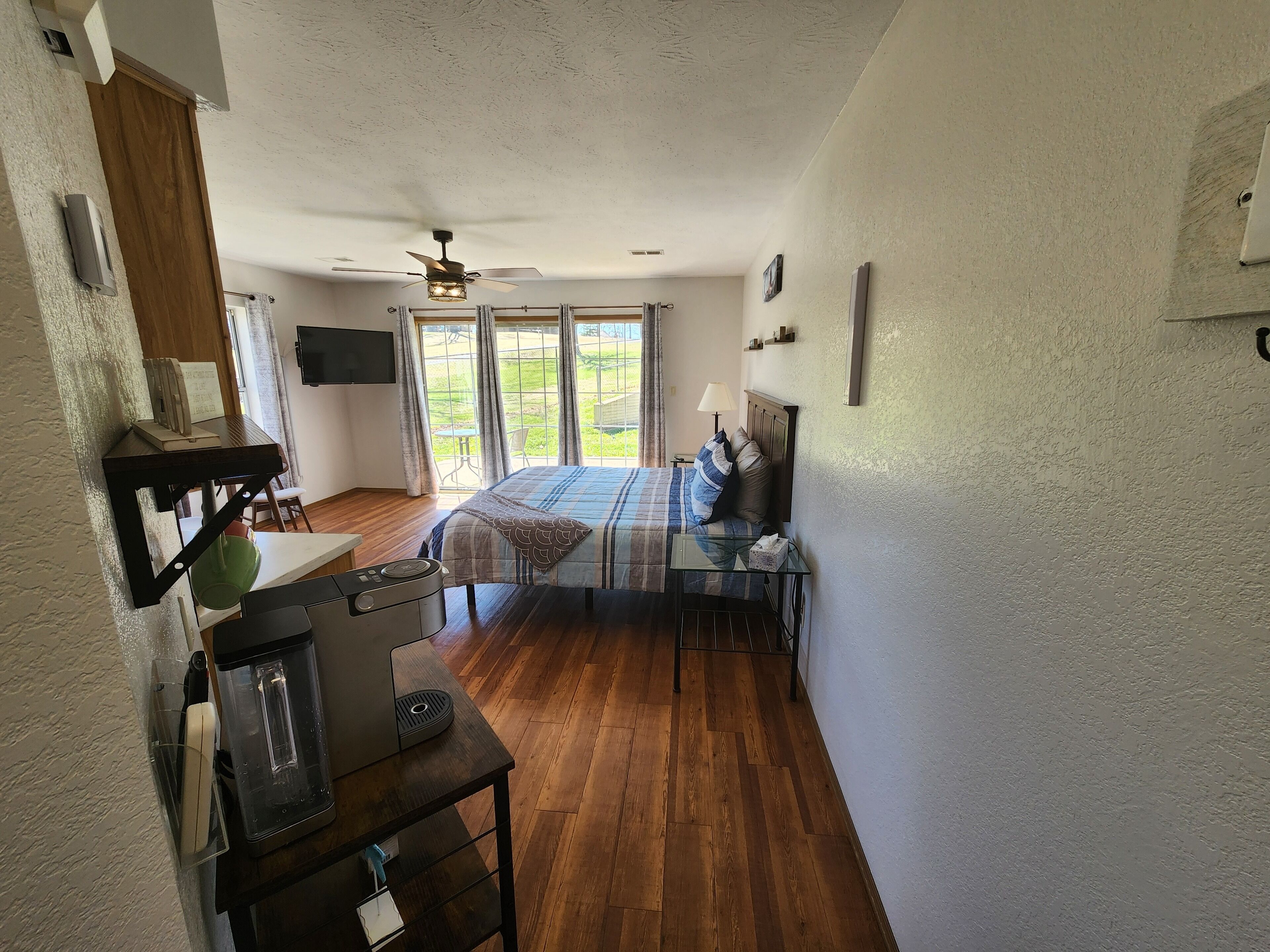A spacious room with a bed, a desk, a coffee machine, and a view of a garden through large windows.