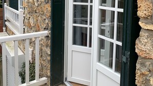 Ein Gebäudeeck mit einer Steinfront, einer weißen Tür mit grünen Fensterläden und einem weißen Balkonläufer.