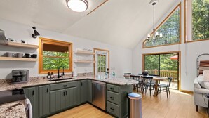 Una cocina moderna con encimeras de granito, gabinetes verdes y lavavajillas integrado. Hay un comedor con mesa y sillas, y un amplio ventanal con vistas al exterior.