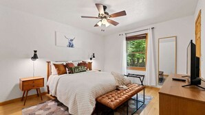 Un dormitorio con cama, mesitas de noche, un banco, ventilador de techo, ventana con vistas y televisor.
