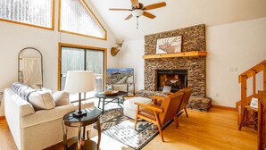 Una sala de estar acogedora con chimenea de piedra, ventilador de techo y ventana grande con vistas a un paisaje nevado.