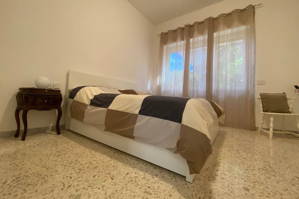A bedroom with a bed, a nightstand, a chair, and a window with curtains.