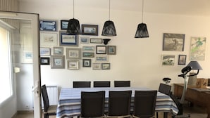 Un coin repas avec une table dressée pour six personnes, entourée de chaises en osier, sous des suspensions noires, et des œuvres d’art encadrées ornant les murs.