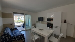 Un espace de vie compact comprenant un canapé, une table à manger avec des chaises, un coin cuisine et une porte-fenêtre coulissante donnant sur un espace extérieur.