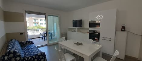 Un espace de vie compact comprenant un canapé, une table à manger avec des chaises, un coin cuisine et une porte-fenêtre coulissante donnant sur un espace extérieur.