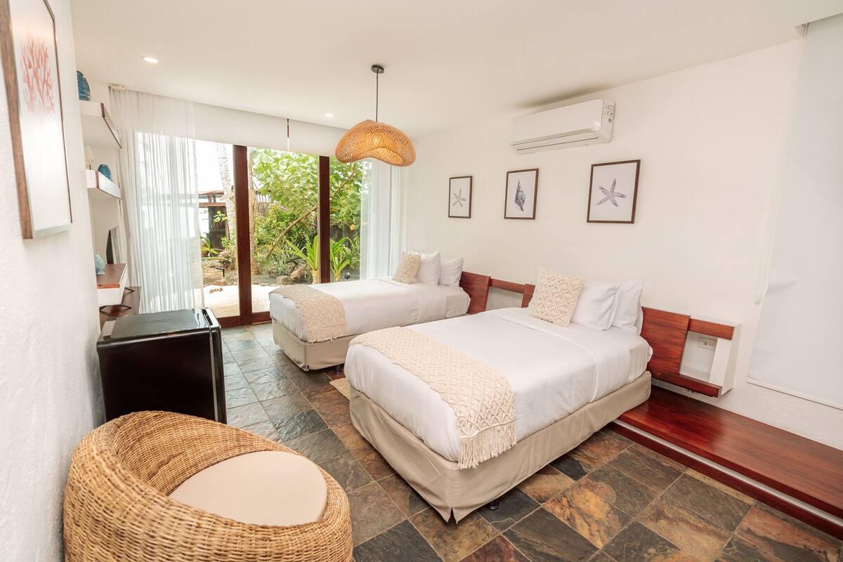 A hotel room with two beds, a wicker chair, a large window with curtains, and a view of greenery outside.