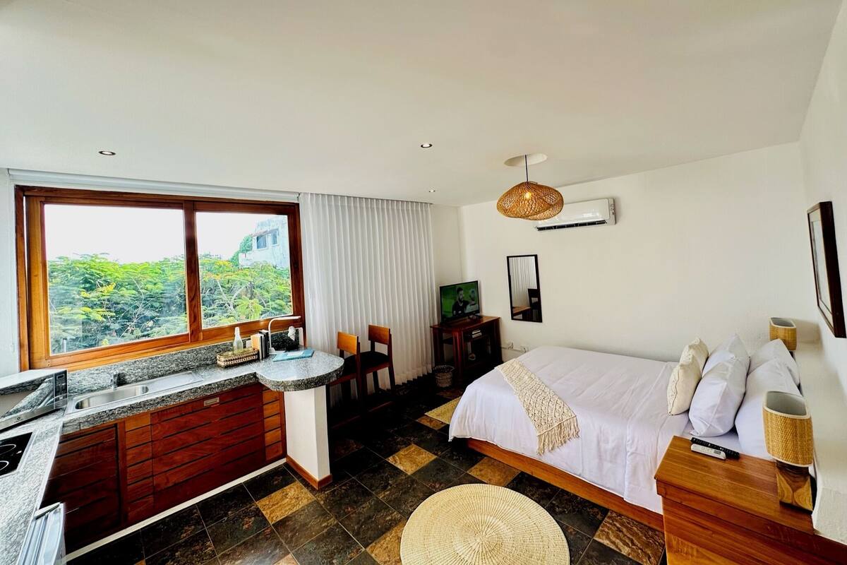A modern hotel room with a kitchen area, a large bed, and a view of greenery outside.