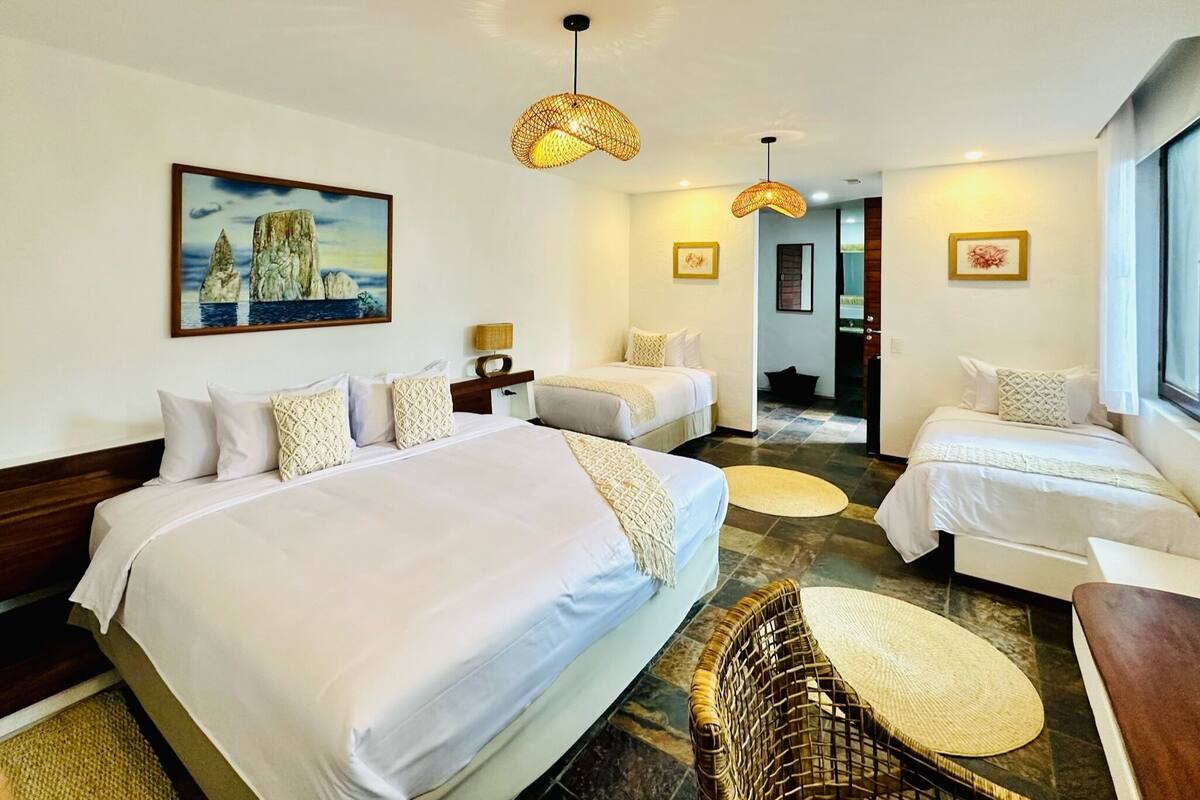 A hotel room with two beds, a wicker chair, and a painting of a coastal scene.