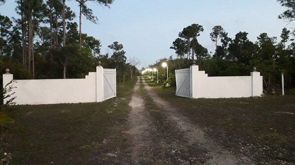 A paved pathway leading to a gated entrance with white walls and metal gates, surrounded by trees and illuminated by street lamps. - PARADISE CREEK COTTAGES (Andros Town)