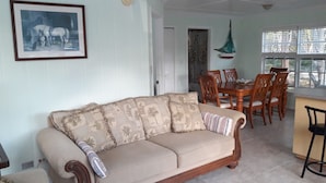 A living room with a sofa, a dining table with chairs, and a kitchen area with a window. - PARADISE CREEK COTTAGES (Andros Town)