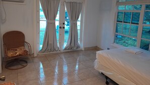 A hotel room with a bed, a chair, a ceiling fan, a window with a view of greenery, and an exit sign. - PARADISE CREEK COTTAGES (Andros Town)