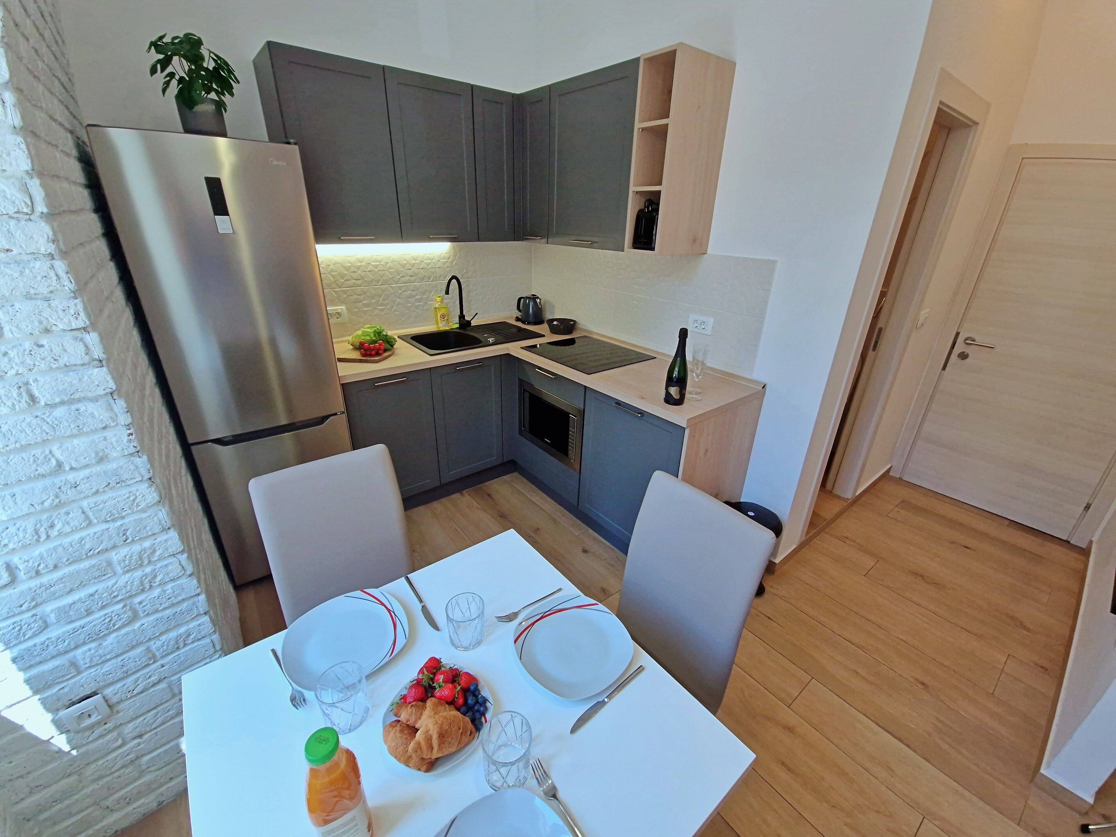 A compact kitchen with grey cabinets, a stainless steel refrigerator, a sink, and a dining table set for two with plates, cutlery, and a bottle of orange juice.