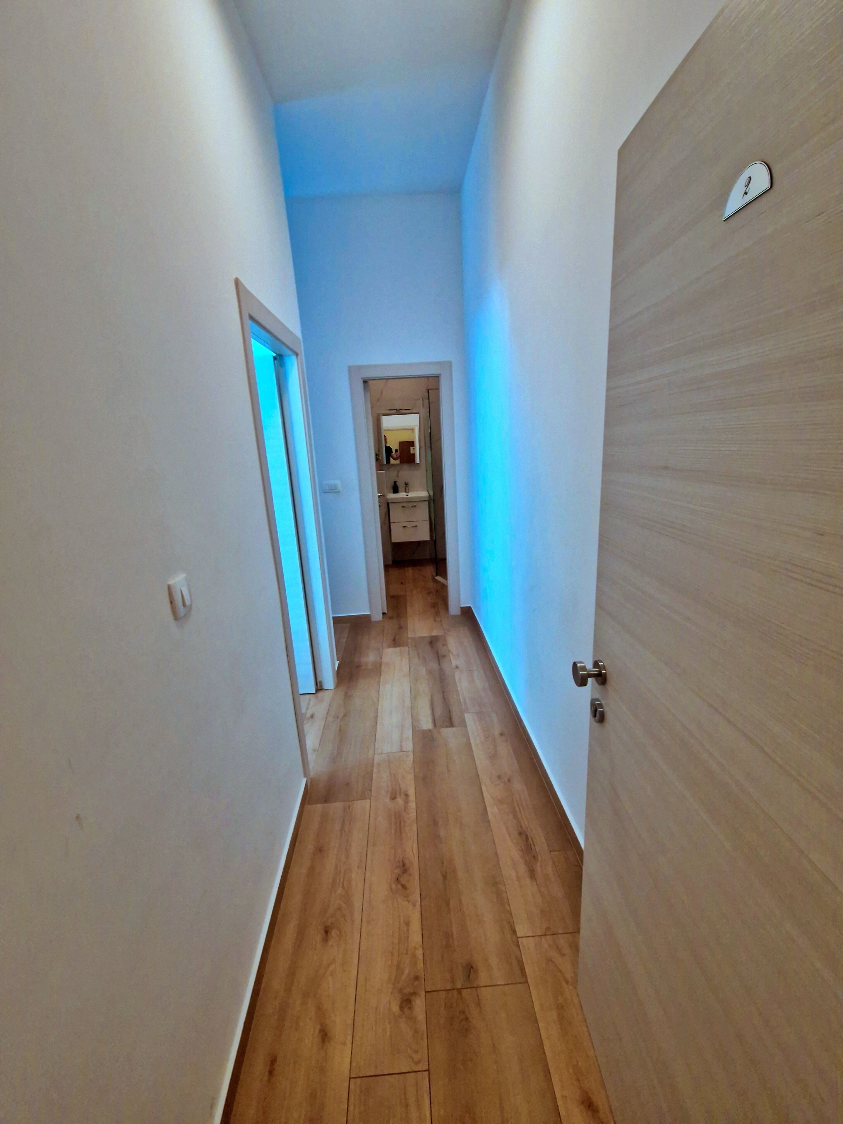 Un couloir étroit, doté d’un plancher en bois et de murs blancs, mène à une salle de bain équipée d’un miroir et d’un lavabo.