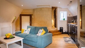 Ein gemütliches Wohnzimmer mit einem blaugrünen Sofa, einem kleinen weißen Tisch mit Früchten und einer Küchenzeile mit modernen Geräten.
