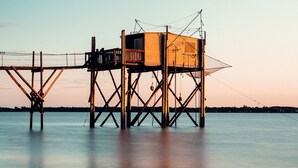 A wooden structure with a platform and a small enclosed area, likely a lookout or observation point, situated over water with a calm surface and a distant shoreline. - L'escapade Fourasine - Heart of Fouras (Fouras)