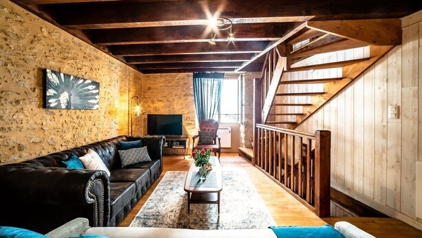 Een rustiek interieur met zichtbare houten balken, een stenen muur, een leren bank in zwart leer, een houten salontafel en een trap.