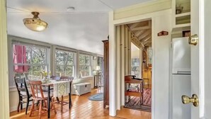 A spacious room with wooden flooring, a dining area with chairs and a table, a ceiling fan, and a view through large windows.