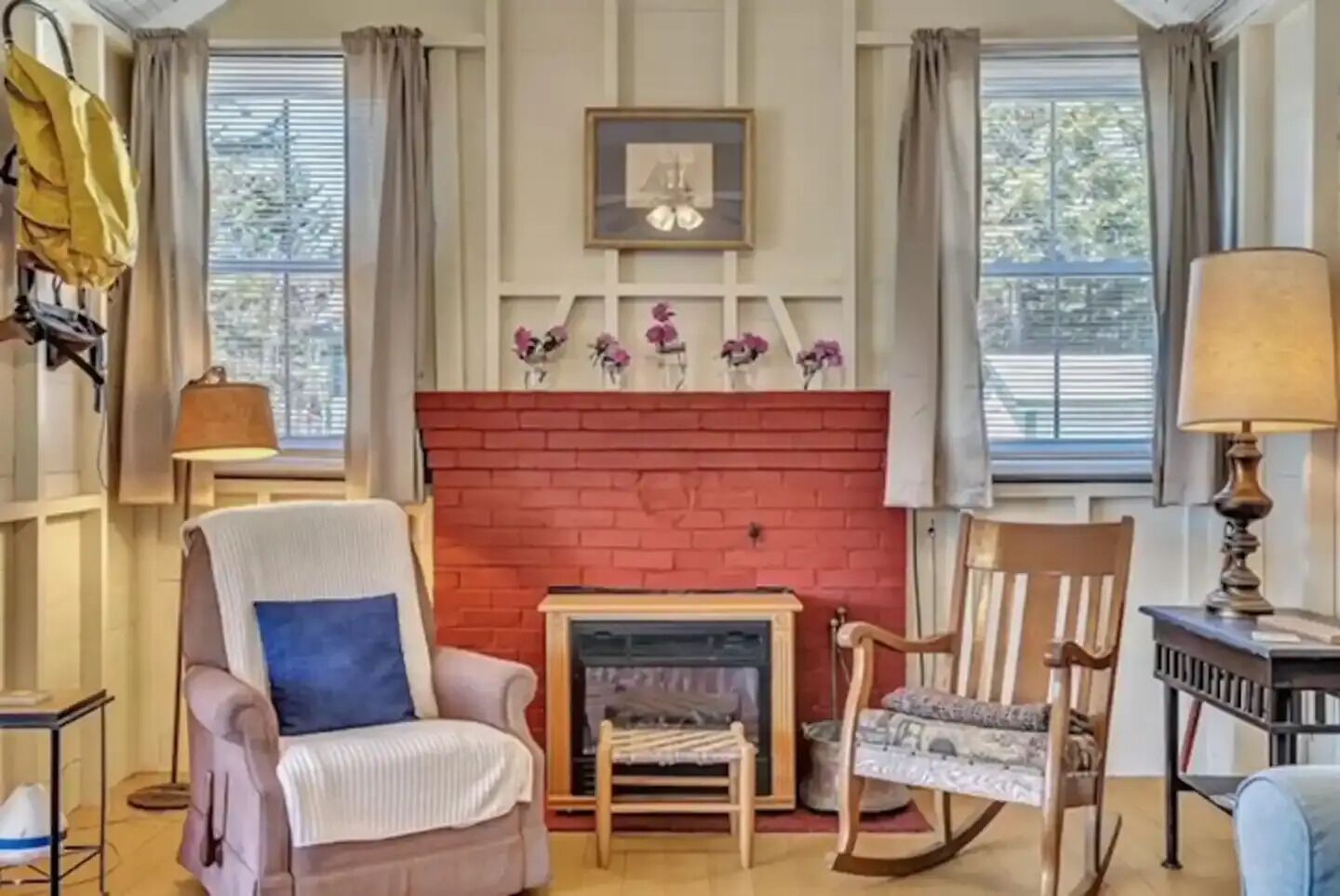 Una sala de estar acogedora con una chimenea de ladrillo rojo, dos sillones, una mesa y una lámpara.