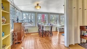 A spacious room with wooden flooring, a dining table with chairs, a bookshelf, and large windows allowing natural light.