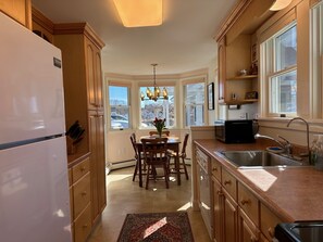 A kitchen with wooden cabinets, a refrigerator, a dining table with chairs, a chandelier, and large windows.