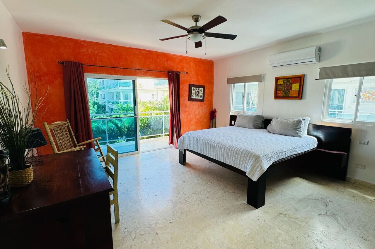 Une chambre à coucher avec un lit, un ventilateur de plafond, une fenêtre avec des rideaux et une vue sur la verdure à l’extérieur.