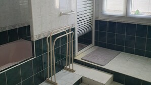 Une salle de bain avec un carrelage au sol, un espace douche et une fenêtre équipée de stores.