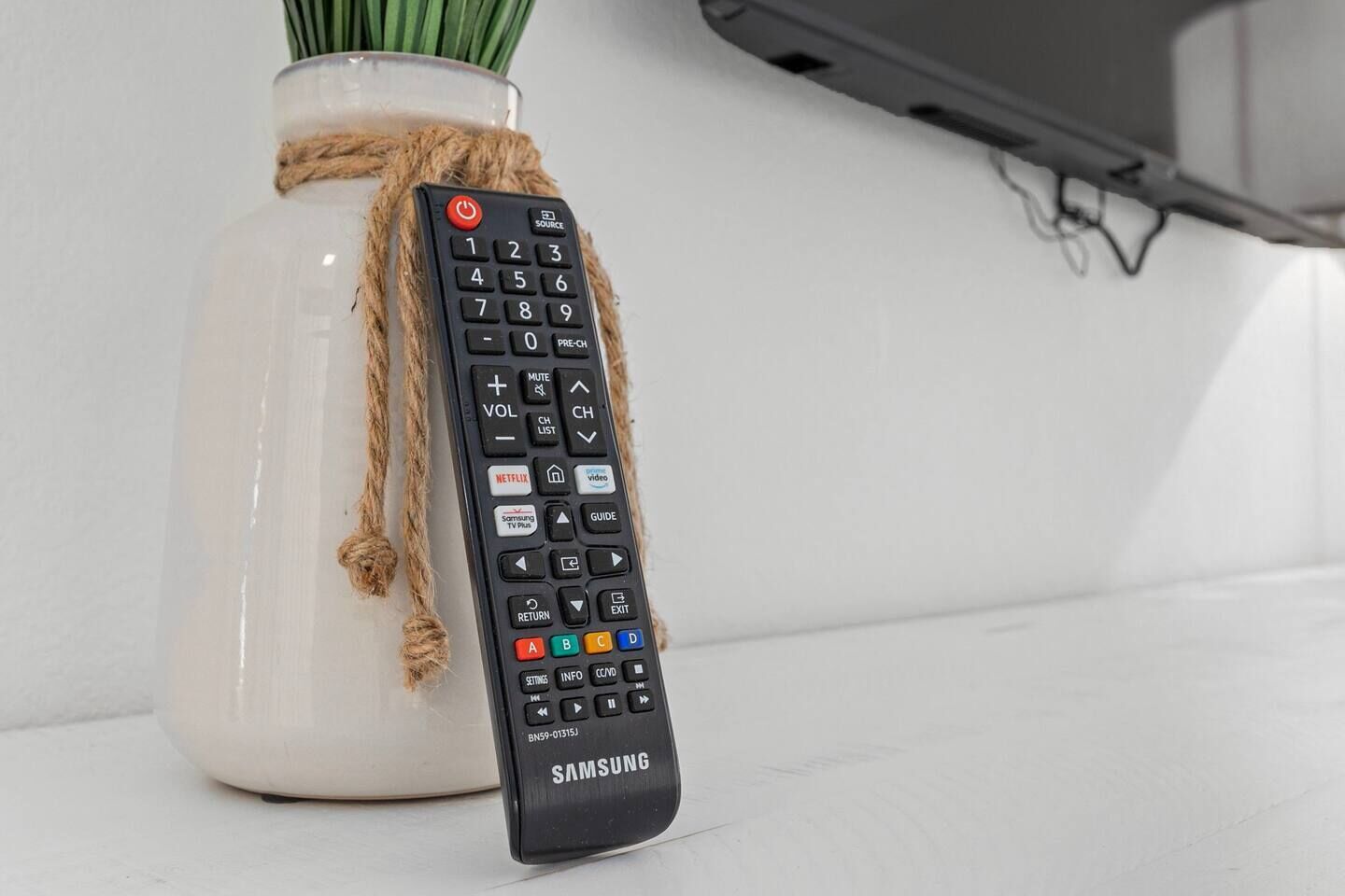 Un telecomando nero Samsung legato con una corda a un vaso bianco su una superficie bianca.