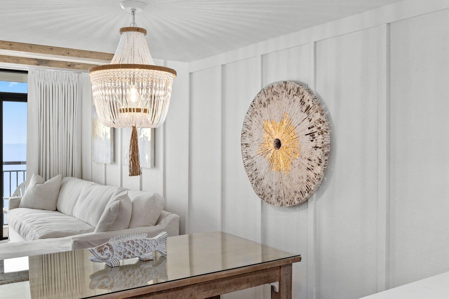 A modern living room with a white sofa, a glass coffee table, a chandelier, and a decorative wall piece.